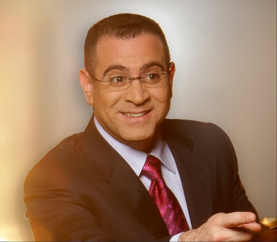 תמונת מופע: אלי יצפאן עם חליפה ומשקפיים מחייך ומצביע קדימה