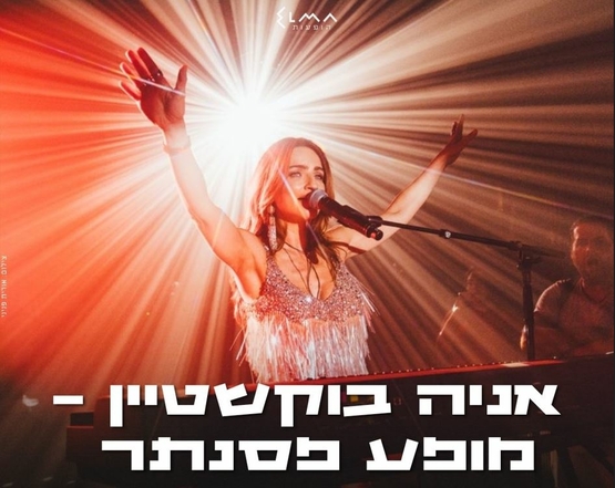 תמונת מופע: אניה בוקשטיין שרה למיקרופון על הבמה ומרימה ידיים